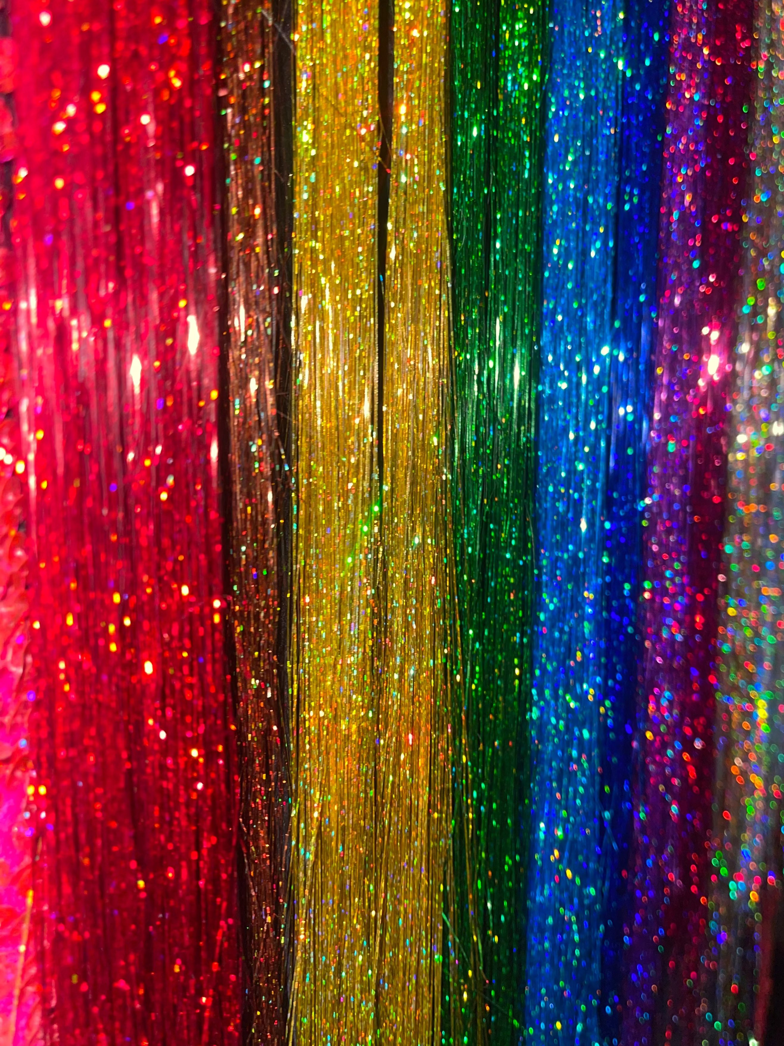 A close-up of vertical strands of glittery tinsel or fringe in a rainbow color sequence, including red, orange, yellow, green, blue, and purple, hanging against a dark background. Hair tinsel parties available