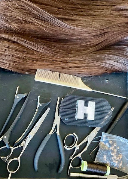 A hair extension kit laid out on a dark tray, featuring a large bundle of medium brown hair extensions, a beige comb, metal and black styling tools, a small black hair clip with a white H logo, a spool of dark thread, and a clear bag of small installation beads.